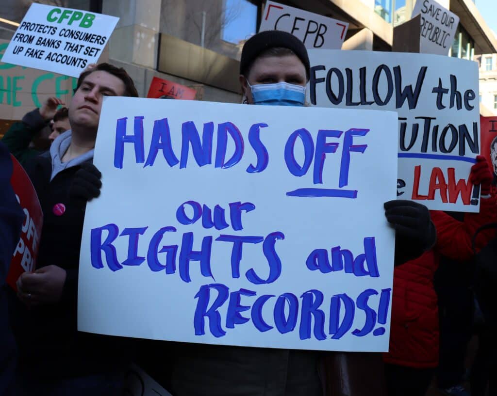 Protestor outside the Consumer Financial Protection Bureau holding a sign that reads "Hands Off our Rights and Records"